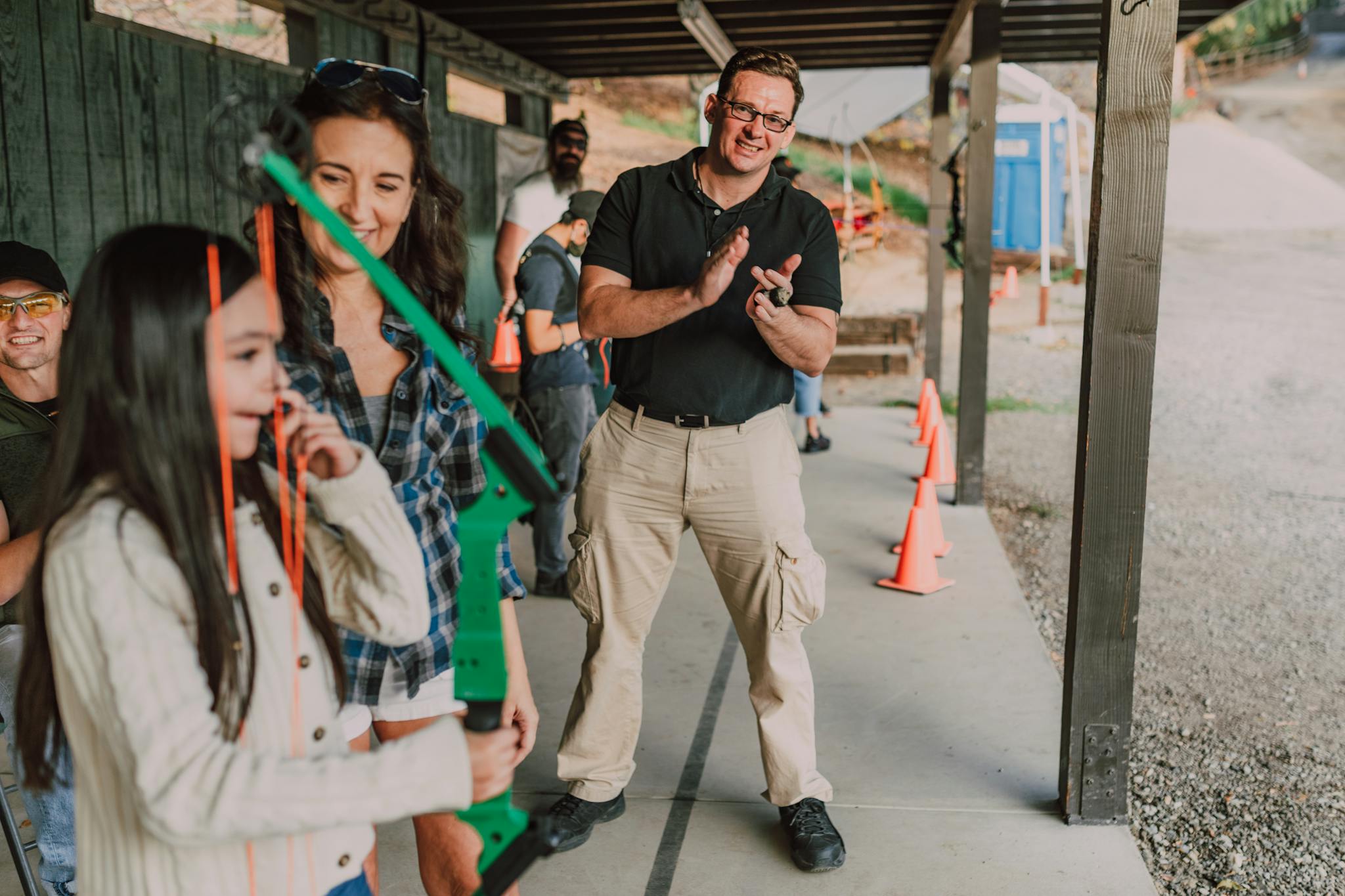 Young girl learning archery outdoors with guidance and support from adults.